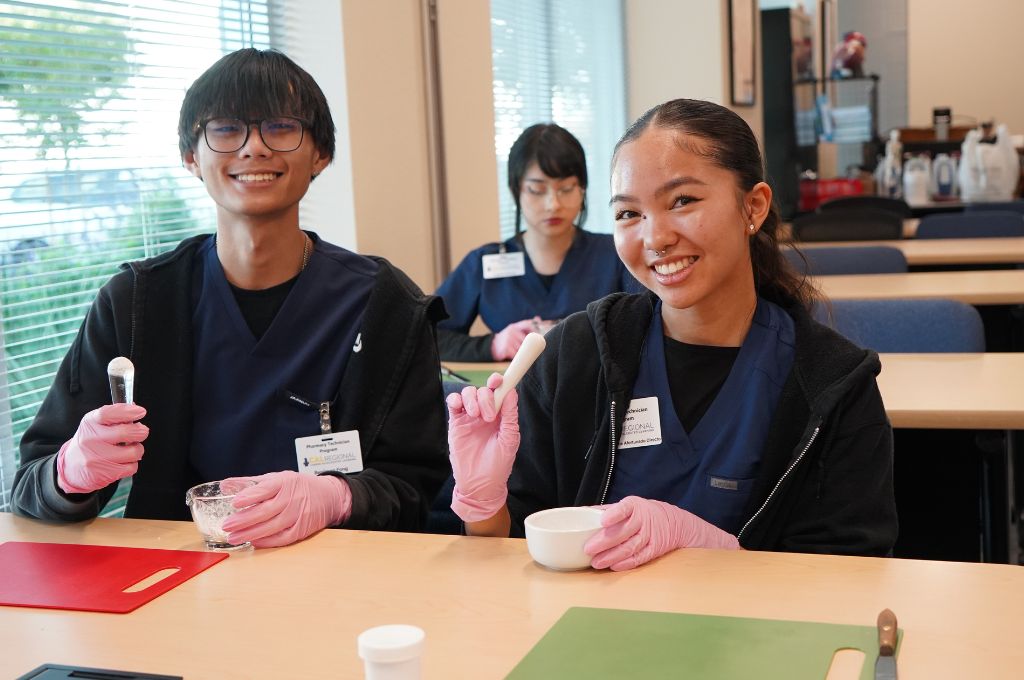 Pharmacy technician students in Texas practicing compounding techniques during a certification training course. They are wearing navy scrubs and pink gloves while using a mortar and pestle to mix ingredients in a classroom setting.