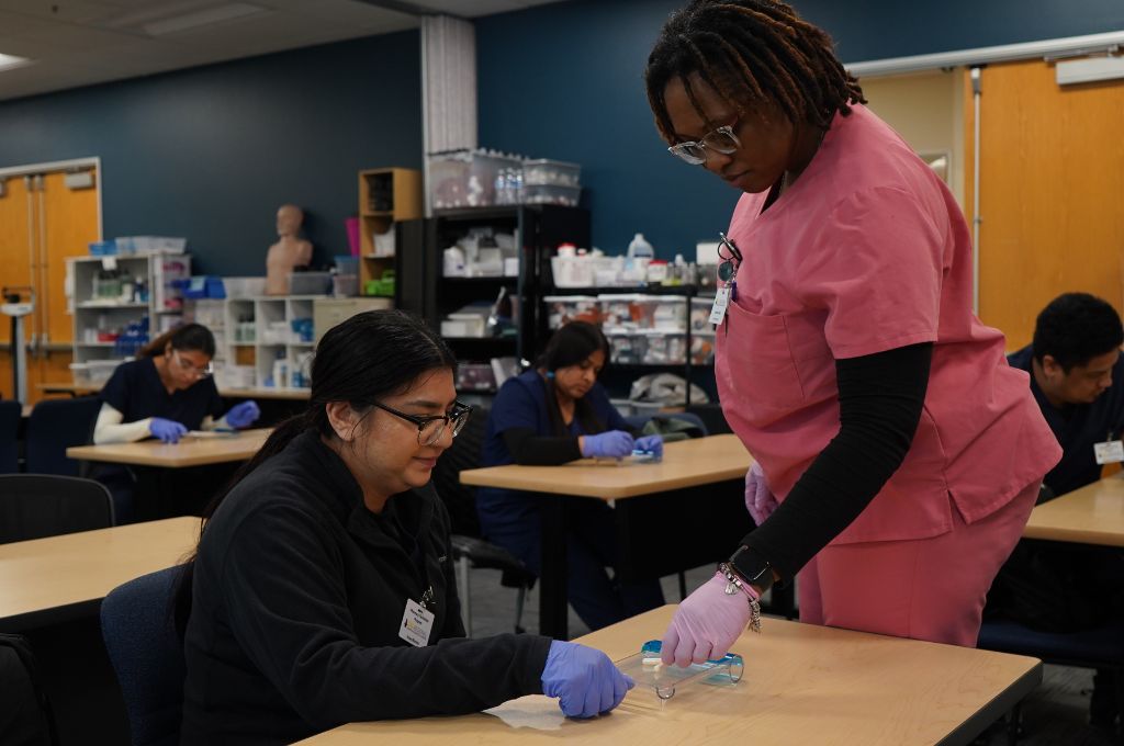 Pharmacy technician students in Texas participating in a hands-on certification training session. An instructor in pink scrubs is assisting a student in black scrubs with a pharmaceutical preparation exercise in a classroom equipped with medical supplies.