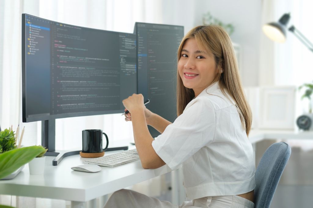 A professional woman working at a clean desk setup with dual monitors and code on screen, smiling while working from home, suggesting flexibility in medical coding careers.